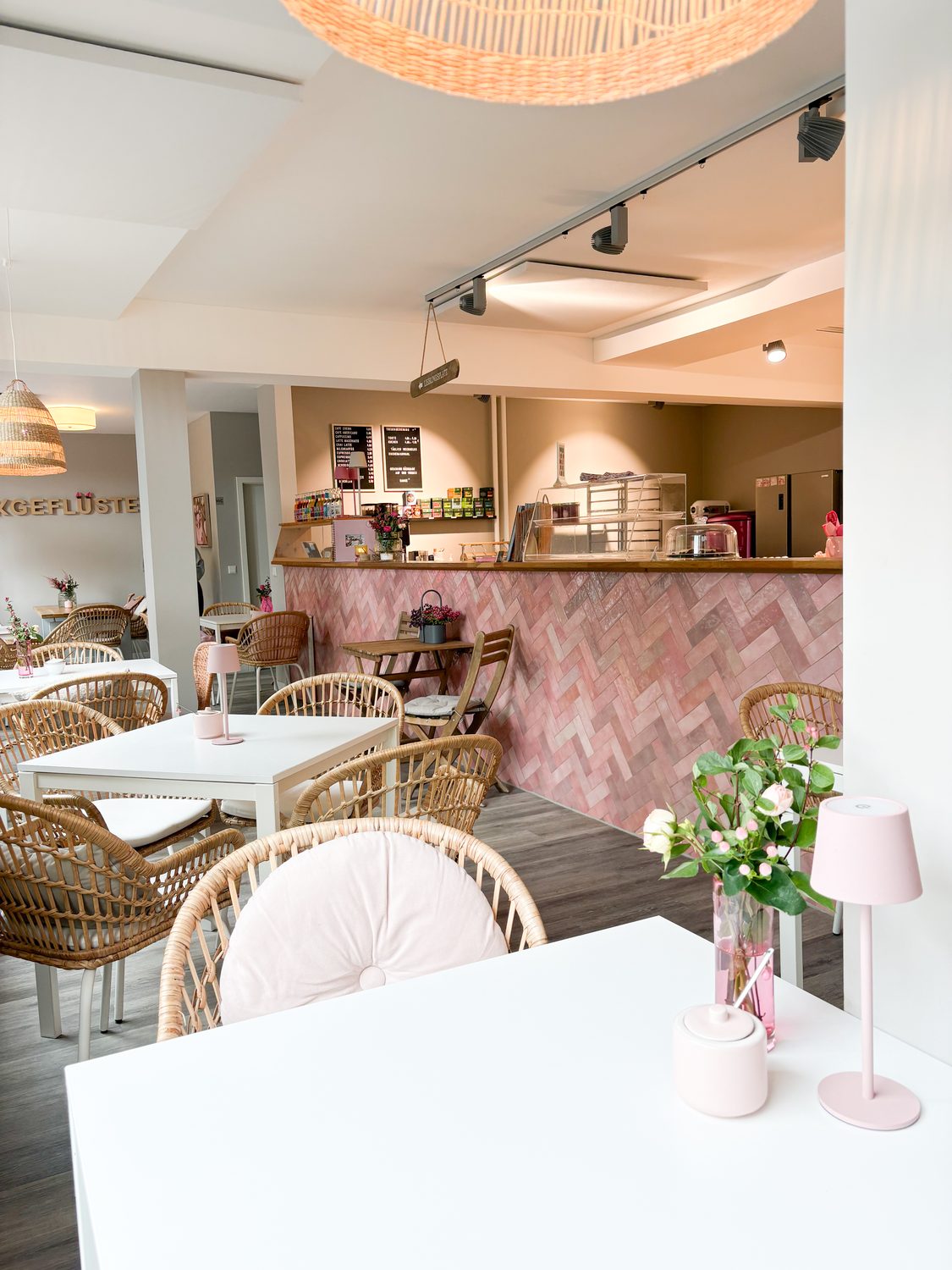 Heller Gastraum im Café Backgeflüster mit Rattanstühlen, weißen Tischen und rosa Tresen